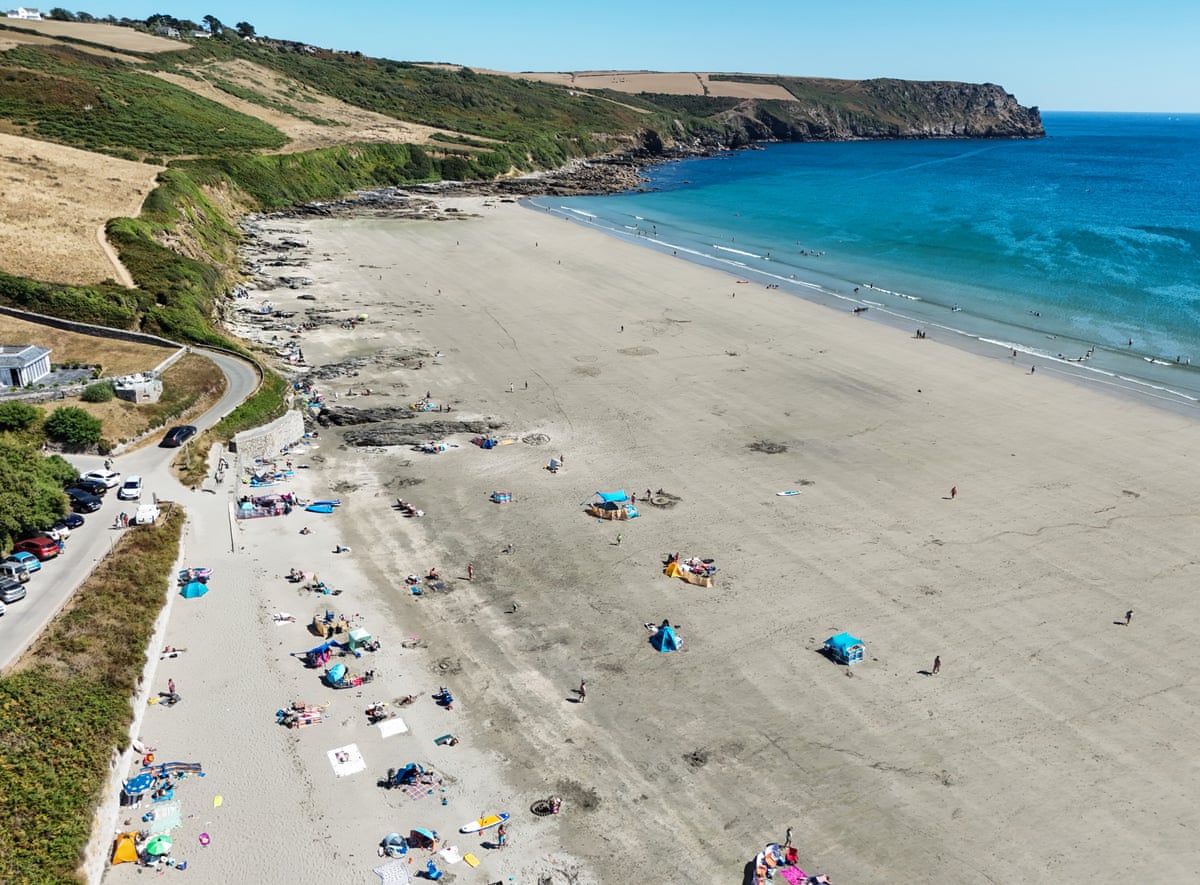 An aerial view of a beach in Cornwall, with blue water and people laid out under umbrellas on the sand.