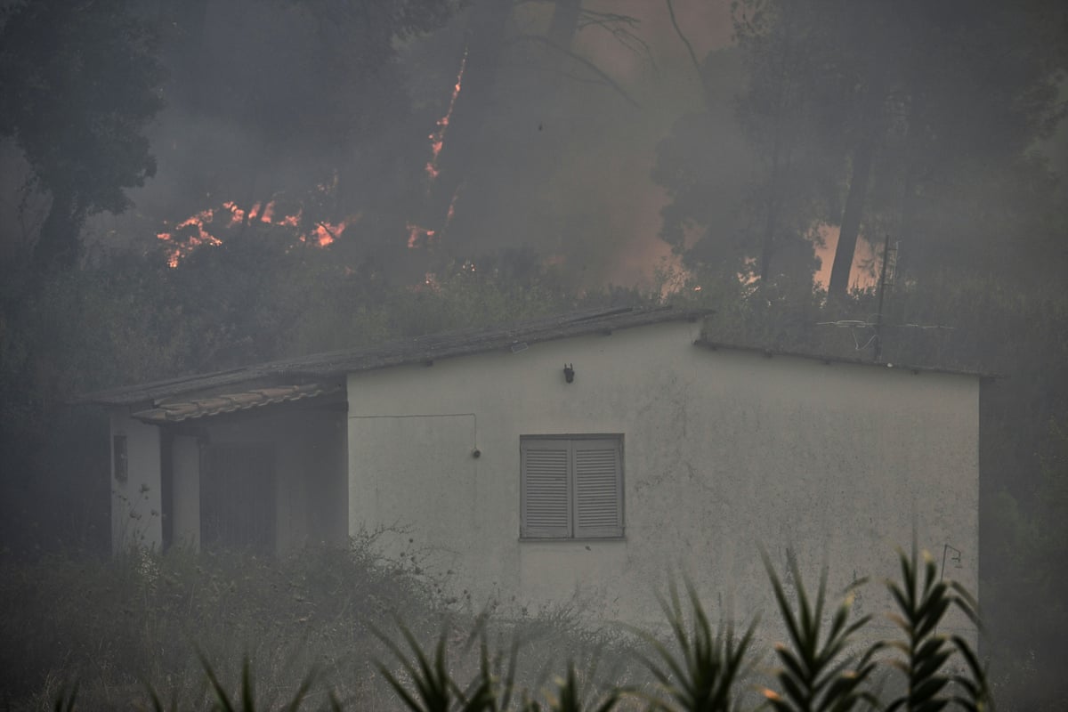 Smoke fills the air with flames in the distance encroaching on a small, white house among trees.