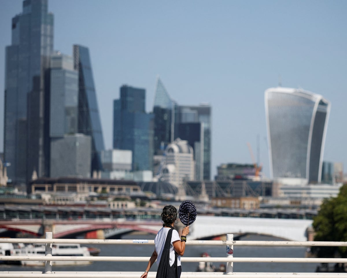 A woman holding a fan walks across Waterloo Bridge, with the City of London financial district in the background, during a heatwave in London.