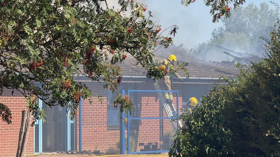 A fire officer is half-way up a ladder leant against a one-storey building wall and roof. Another officer is on the ground looking towards the person on the ladder. Some double doors are open and there is shrubbery and a tree in the front of the building.