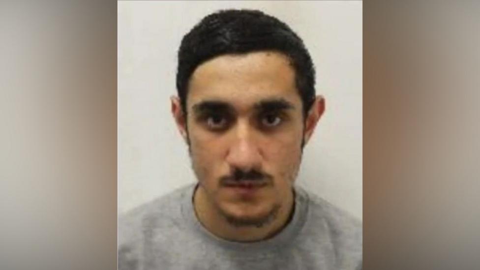 Head-and-shoulders custody photograph of a person with short dark hair wearing a grey top, facing the camera against a plain light-coloured background. The image is tightly cropped around the face and shoulders.