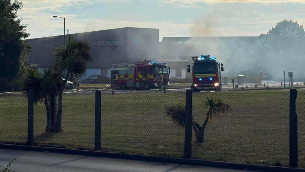 Fire engines outside the fire station. The image is taken through a fence surrounding the site. The smoke has thinned out. Some fire officers can be seen in front of their vehicles.