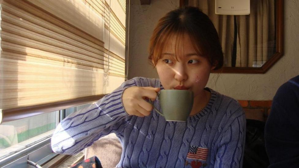 Soomin Yi sipping on a cup of tea while glancing towards a window to her right