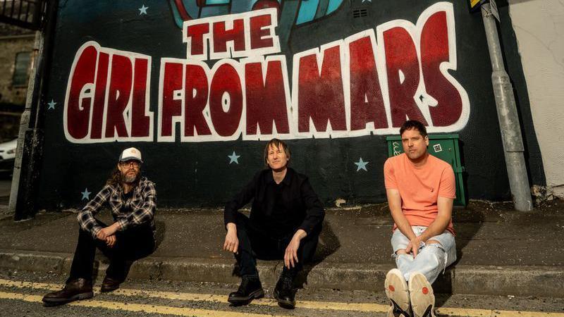 Three men sitting casually on a pavement below a mural which says 'The Girl From Mars' in large red graffiti. They look relaxed.