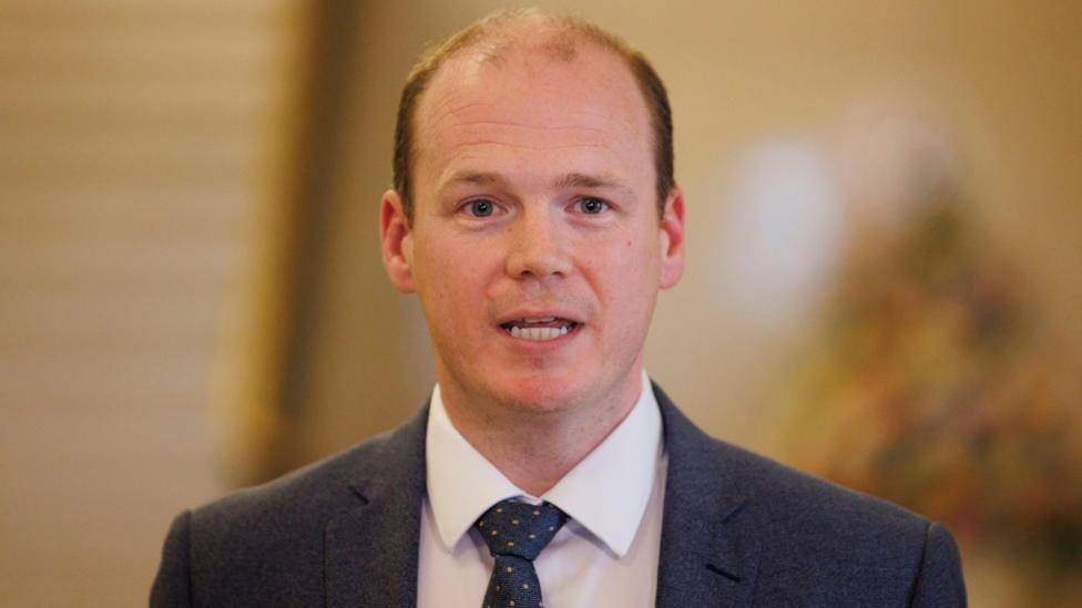 Communities Minister Gordon Lyons pictured during a press conference at Parliament Buildings at Stormont in Belfast. He has thinning brown hair. He is wearing a dark suit, white shirt and navy tie with pink spots.