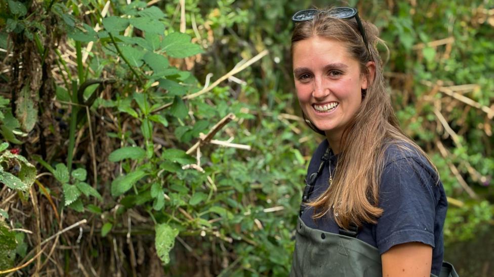 A woman with long brown hair and sunglasses on her head smiles at the camera. She is wearing green waders and a blue t-shirt. She is surrounded by green vegetation.