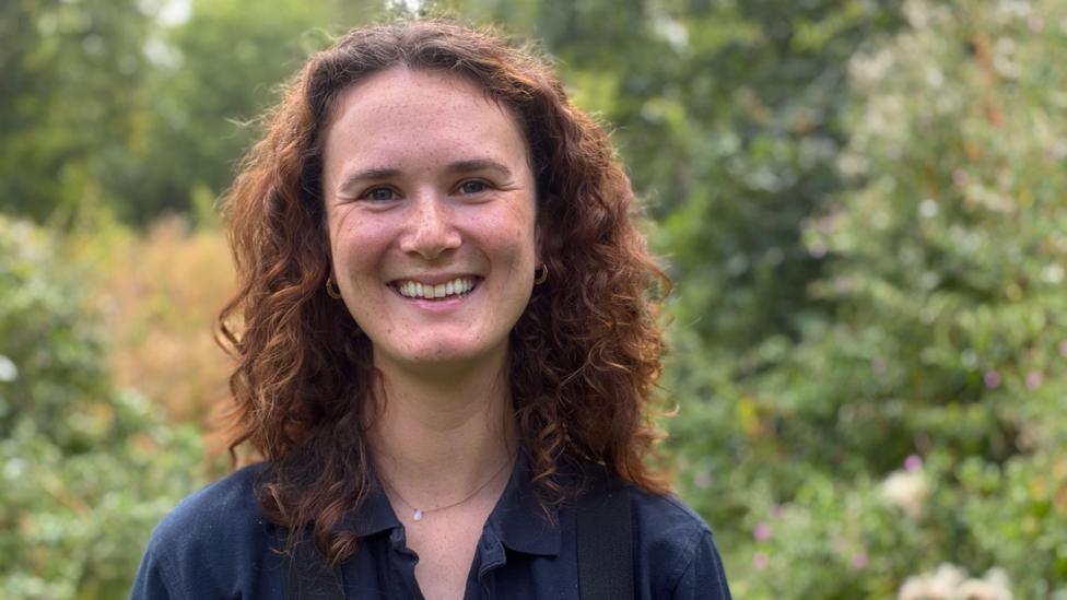 A woman with long curly red/brown hair smiles at the camera. She has a blue collared shirt on and there is a lot of green vegetation in the background.