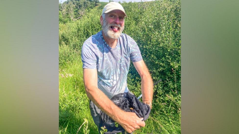 Dave Wainwright looking up at the camera and smiling while holding the small moth within a black net. He is wearing a cream cap and has a white beard. He is wearing a faded blue t-shirt with what looks like a butterfly design on it. He is standing in a field with long grass behind him.