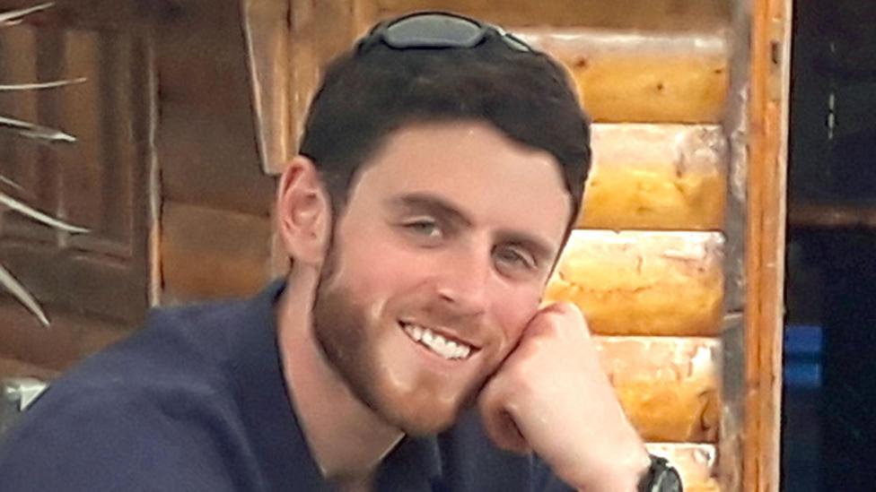 a man with short brown hair and a slight beard wearing a shirt and smiling at the camera.