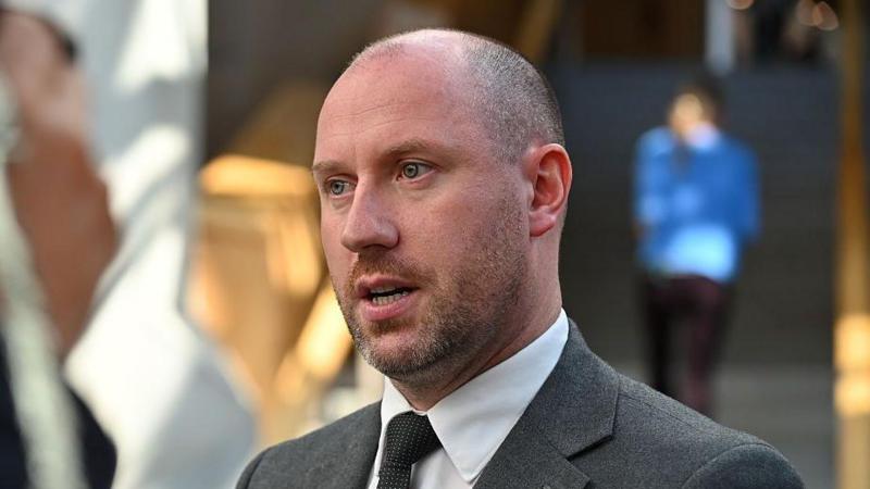 a man with close-cropped hair and beard is wearing a grey suit, white shirt and black tie