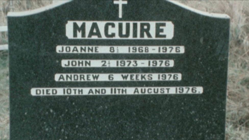 A gravestone with the nhame 'MAGUIRE' engraved in black capital letter into a surrounding block of white. The names 'JOANNE', 'JOHN' AND 'ANDREW' are also engraved in a similar way. At the top is a cross and beyond the white is granite. 