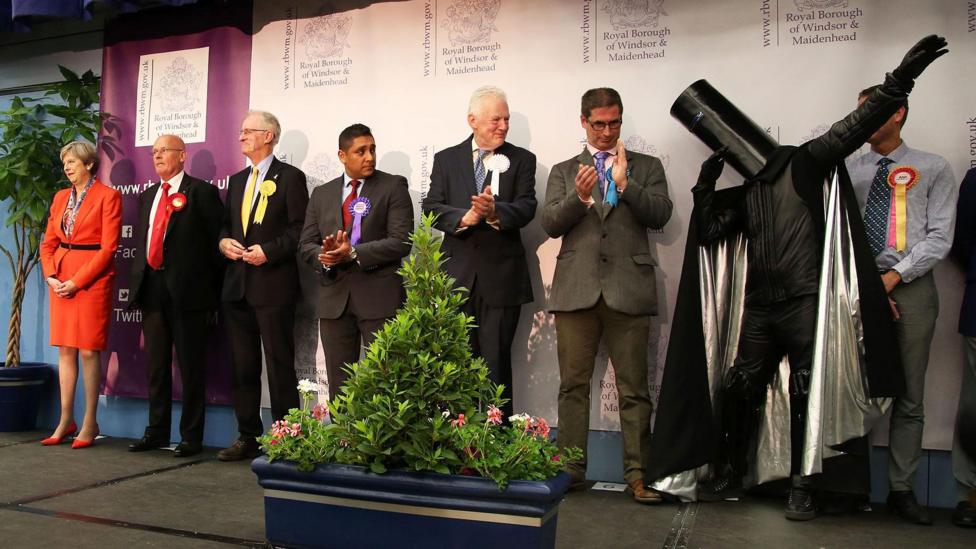 A row of candidates can be seen on stage at the election count. Theresa May is on the far left of at least nine candidates on stage. She wears a bright red skirt suit and matching high heels, with her grey hair in a neat bob. She has her hands clasped on front of her and is smiling wryly. Lord Buckethead is on the far left of the row of candidates and is doing a dab. He wears a large black bucket on his head, with black armour and a black cape with a sliver lining.