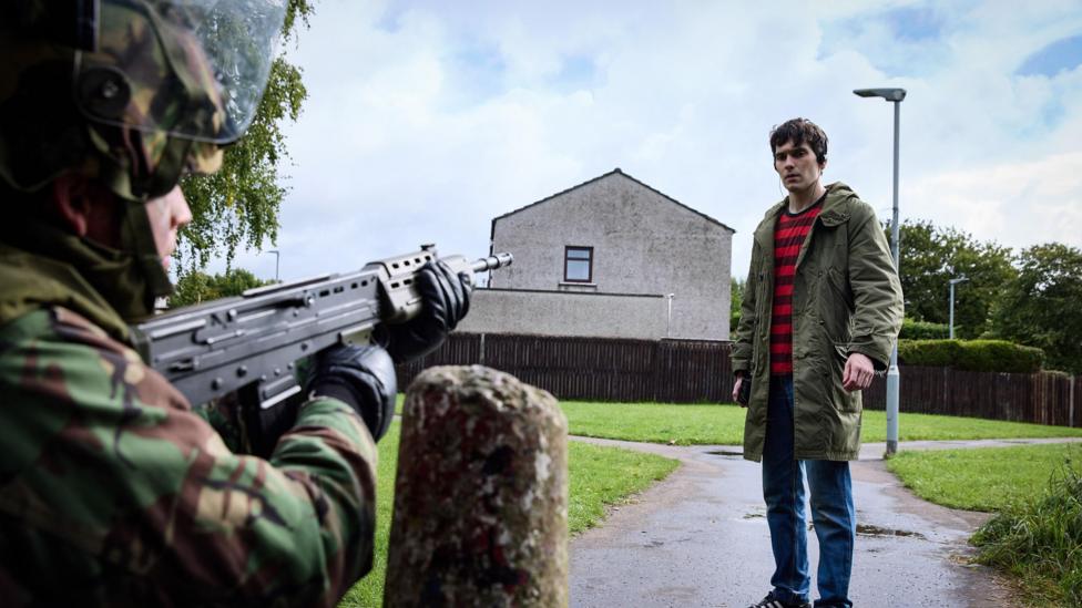 A crouched solider point his gun at a young man in a green parka coat. Behind him is a terrace house, a lamppost, and a path that forks through a green.
