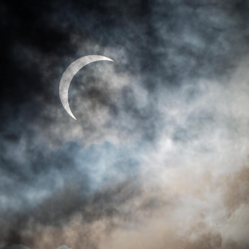 A thin, bright crescent of the Sun shines through layers of dark grey and blue clouds. The scene has a dramatic, moody look, with dense cloud banks along the bottom of the frame.