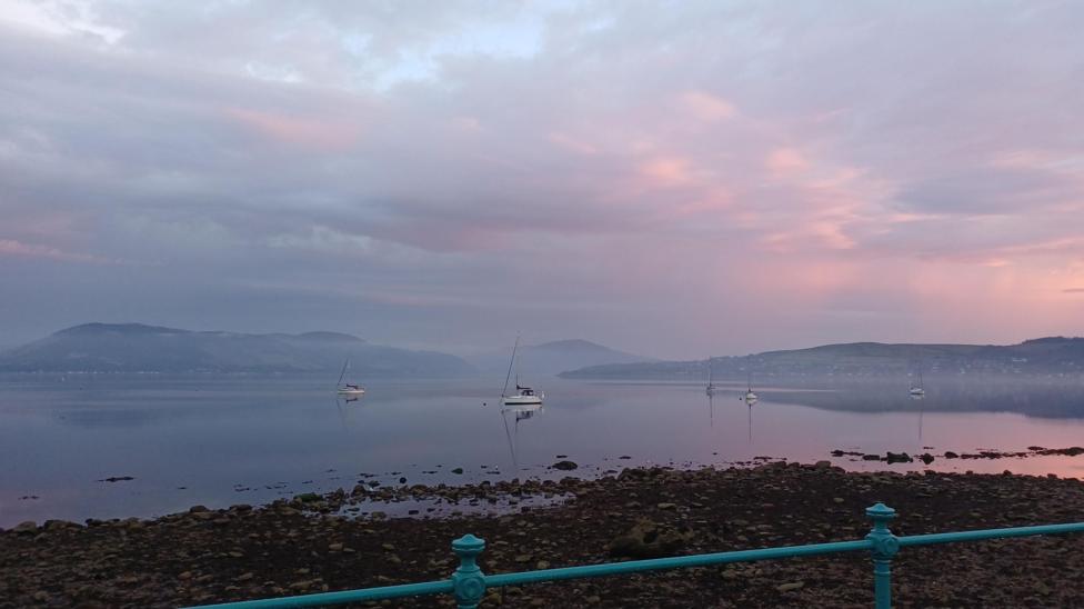 A calm shoreline scene with sailboats anchored on still water under a dramatic sky tinged with pink and purple hues. Mist hangs over the distant hills, while the water reflects the soft evening colours.