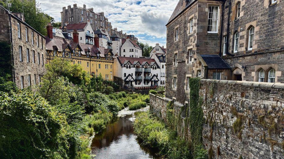 A narrow river winds through a lush green corridor, bordered by stone buildings and traditional timber-framed houses. The scene features historic architecture, ivy-covered walls, and a partly cloudy sky.