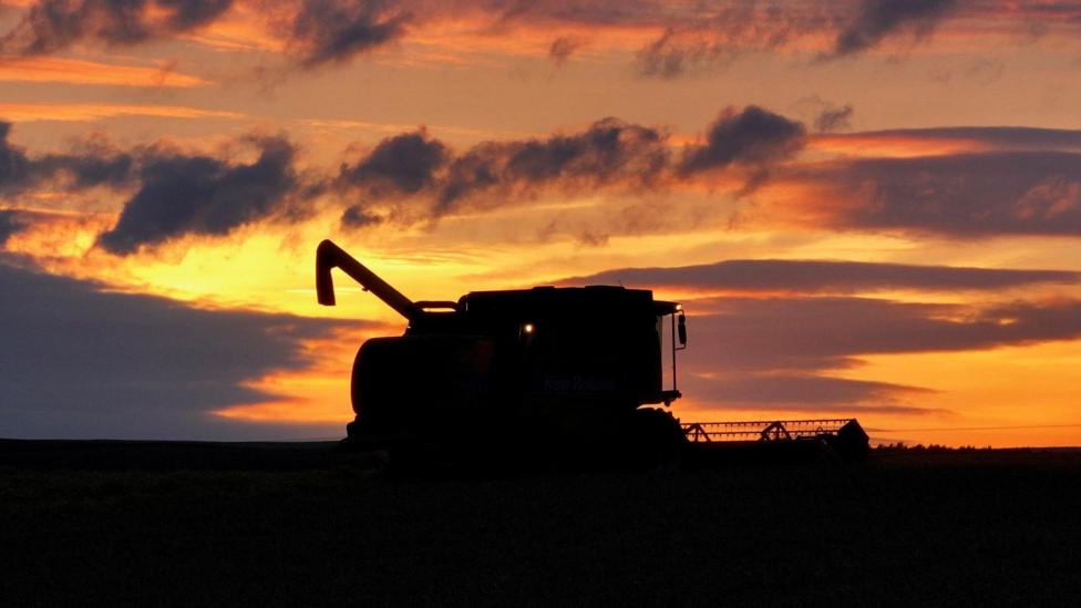 A combine harvester appears in silhouette on an open field, framed by a dramatic sunset. Orange and pink clouds fill the sky, creating a striking contrast with the dark landscape.