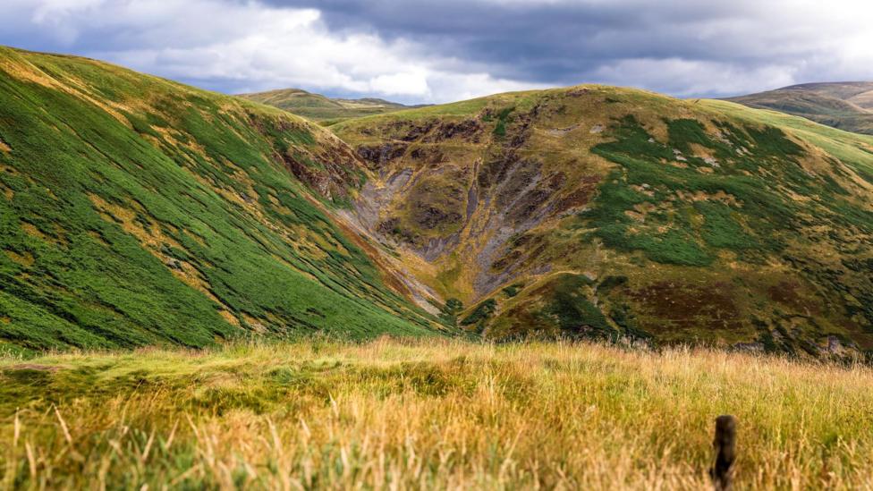 A broad landscape of green and golden hills surrounds a steep-sided valley beneath dramatic, cloud-filled skies.