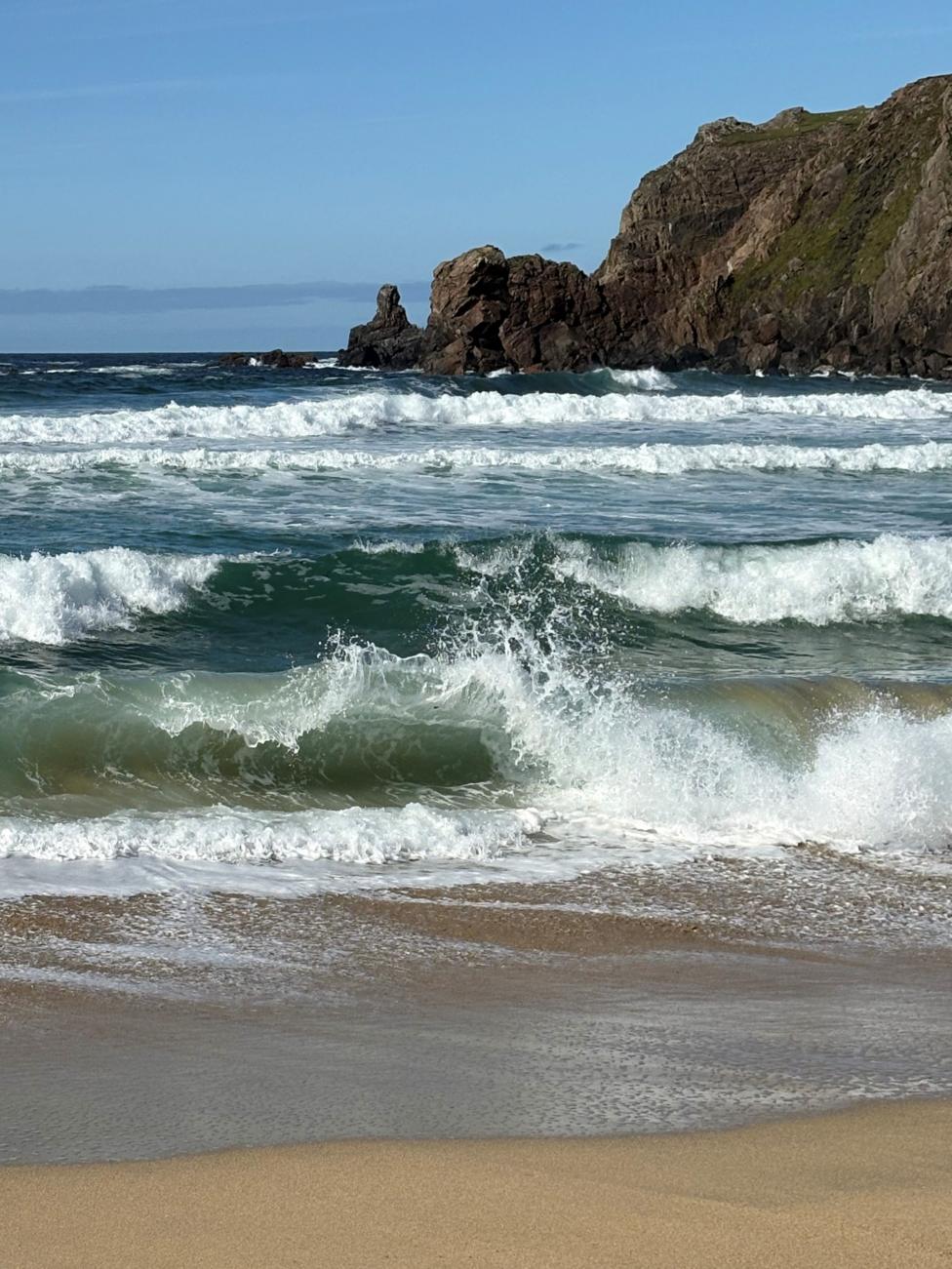 A sandy beach is bordered by churning waves rolling in from the sea. In the foreground, a breaking wave creates white spray and foam as it reaches the shoreline. Beyond the surf, multiple bands of waves stretch across the water towards a rugged rocky headland