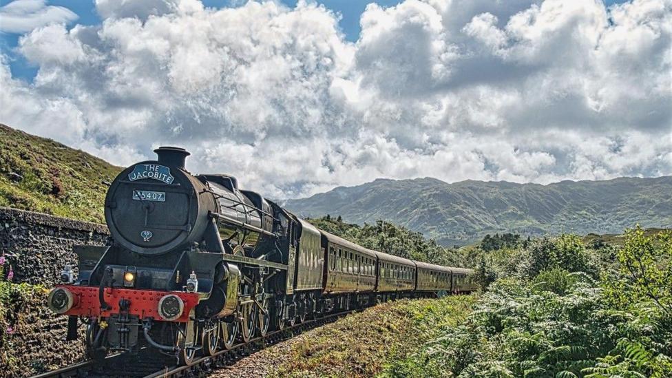 A vintage black steam locomotive pulls passenger carriages along a railway line through a lush green landscape, with mountains and dramatic clouds in the background.