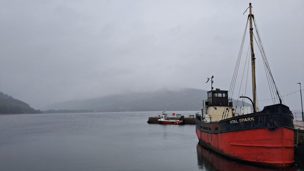 A red-and-black vintage boat named “Vital Spark” is moored beside a calm harbour on a misty day. Low clouds and fog partially obscure the hills in the background. A smaller boat is tied up at a nearby pier, while the still water reflects the larger vessel.