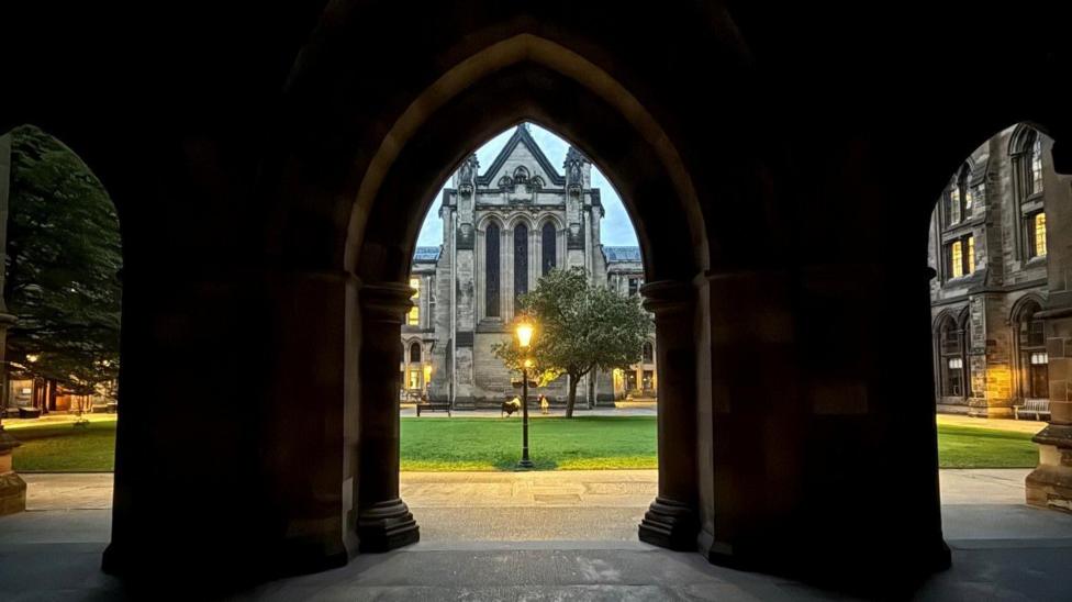 A photograph taken from inside a dark stone archway, looking out onto a courtyard with green grass and a lit lamppost. Framed by Gothic-style architecture, a large stone building stands in the background.