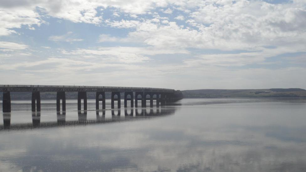 A long bridge stretches across a wide estuary under a partly cloudy sky. The still water mirrors both the bridge and the clouds.