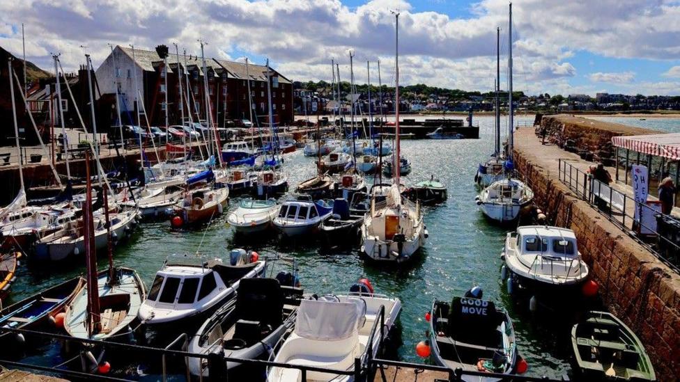 A colourful harbour filled with sailboats and small motorboats moored along stone quays. Bright blue skies and large white clouds overhead add contrast to the bustling waterfront and the buildings lining the harbour.