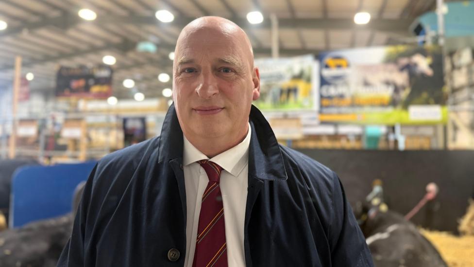 John McLenaghan, a man with shaved head, wearing a blue jacket, white shirt and claret tie. He appears to be in a shed.