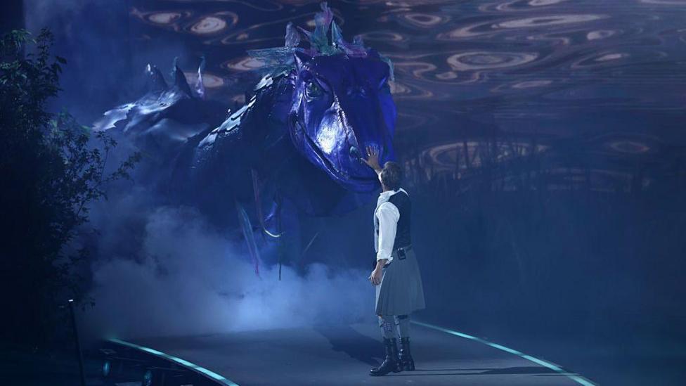 The actor SamHeughan stands onstage, wearing a kilt, and with his back to the camera. He is looking at a giant Loch Ness monster prop that is emerging on stage. It is scaled and coloured purple.