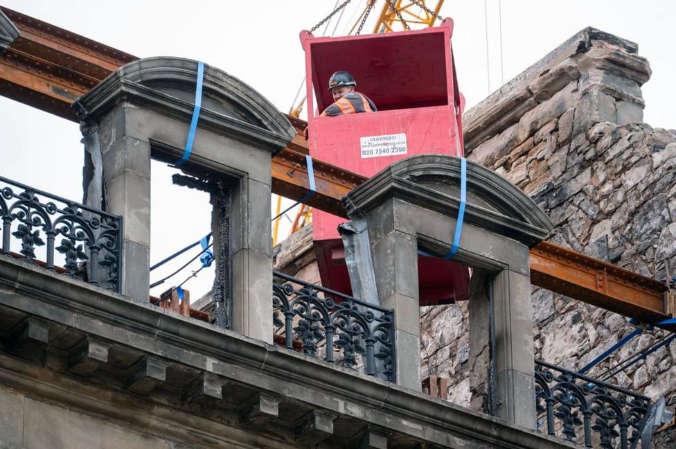 A man looks out of a red suspended crane platform while working on teh damaged facade of an old building