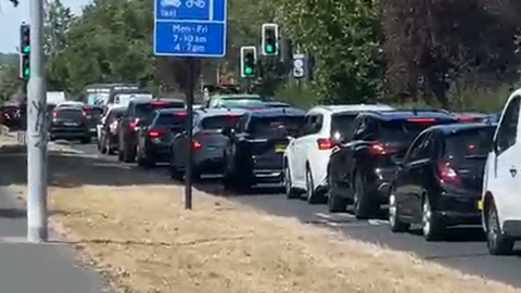 Daytime traffic jam on a road, showing a long queue of cars and a white van stationary near traffic lights, with blue road signs for bus and cycle lanes visible in the foreground.