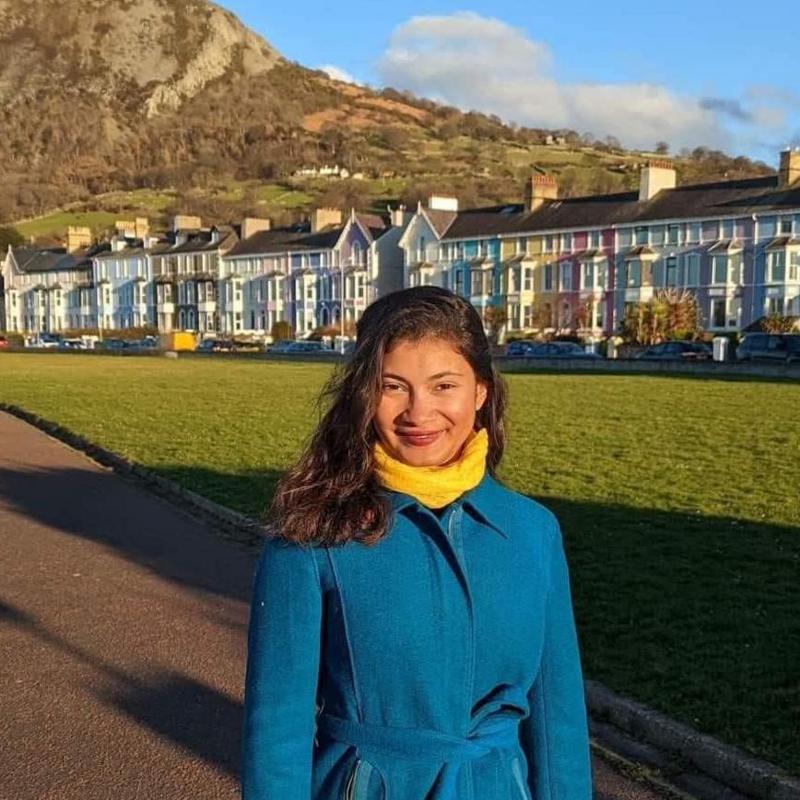 A woman with long dark hair wearing a yellow scarf and blue coat, stood on a path with a patch of grass and row of colourful houses bbehind her. Behind the houses is a mountain.