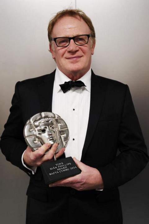 A man with brown hair and glasses, wearing a tuxedo and holding a Bafta Cymru award.
