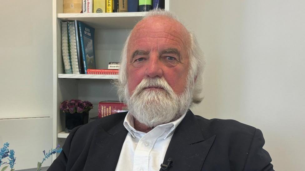A man with white hair and beard, wearing a white shirt and black suit jacket. He is sat down, with a bookcase behind him.