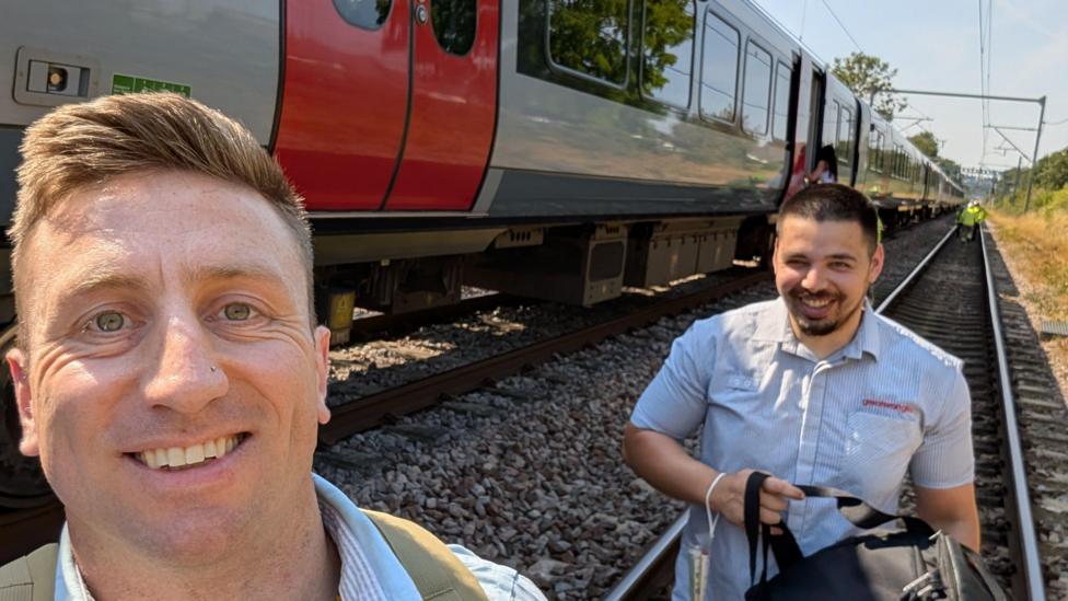 Jon Young has taken a selfie with a member of Greater Anglia staff. Both are walking along the railway line next to the train and wearing pale blue shirts.
