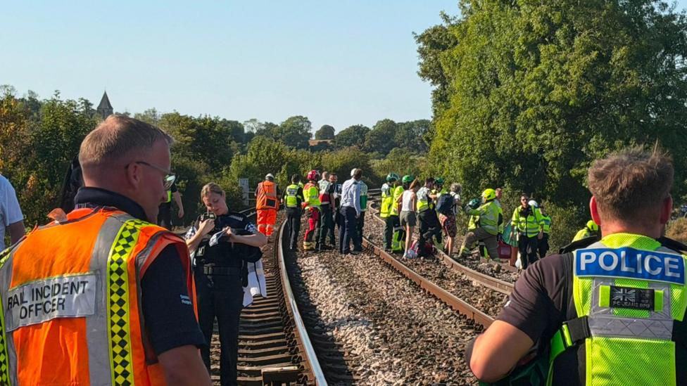 Emergency service personnel, rail staff and passengers stand on a railway line near Lewes in East Sussex following the derailment. Police officers and rail incident officers are visible in the foreground while groups of people gather further along the track during the response to the incident.
