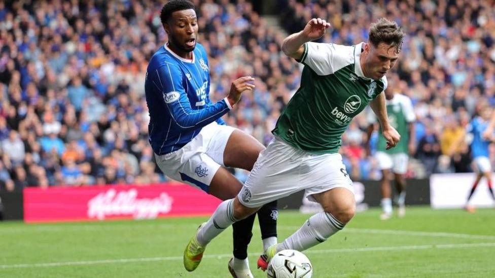 Two football players mid tussle. One is wearing blue and the other green. A football is visible in front of them and a stand of fans behind.