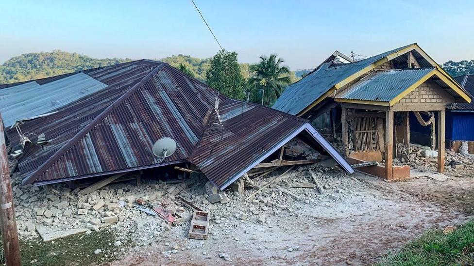 A house has collapsed so that only the roof is visible, lying on the ground.