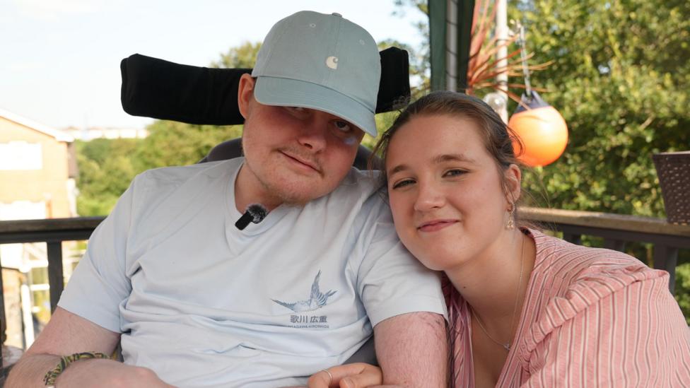 Charlie and Judy smiling at the camera. He is wearing a blue cap and blue t-shirt. Judy is wearing a pink striped dress. They are both in the garden sat down, holding hands.