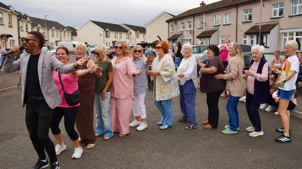 A sharply dressed man is holding a microphone while leading a line of women in a conga.