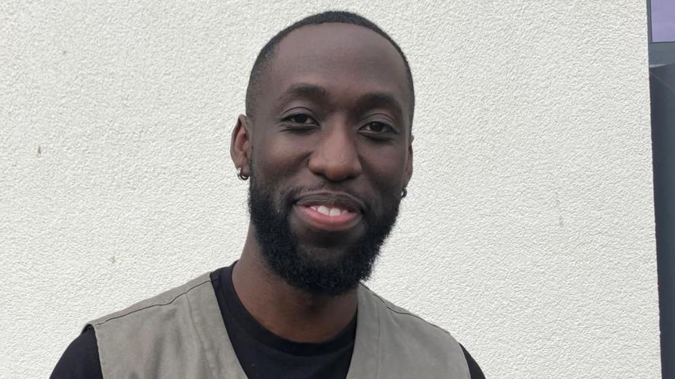 A man in a black t shirt and grey waistcoat stands in front of a grey wall. he has short black hair and beard and is smiling