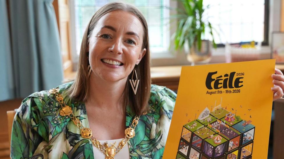 A woman weraing a mayoral chain and a green dress is sat and is smiling. She has long brown hair and is holding a book, with the word feile on its front