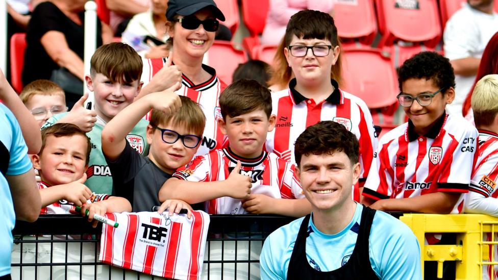 A number of young fans stand in front of a row of stadium seats. in front of them is a footballer. The kids are in football kits.