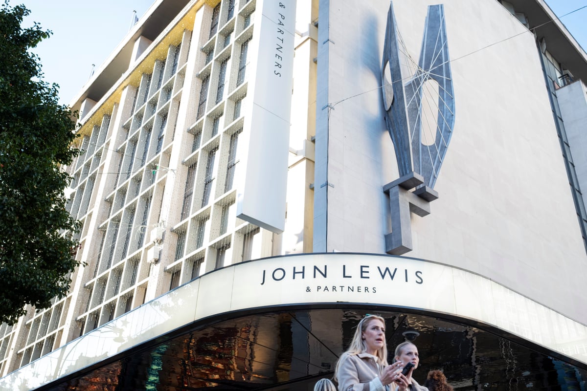 Frontage of the John Lewis store on Oxford Street, London, with an angular modernist sculpture above the doorway that curves around one corner. Two women walk in the foreground and the upper storeys’ white facade with recessed windows stretches along the street.
