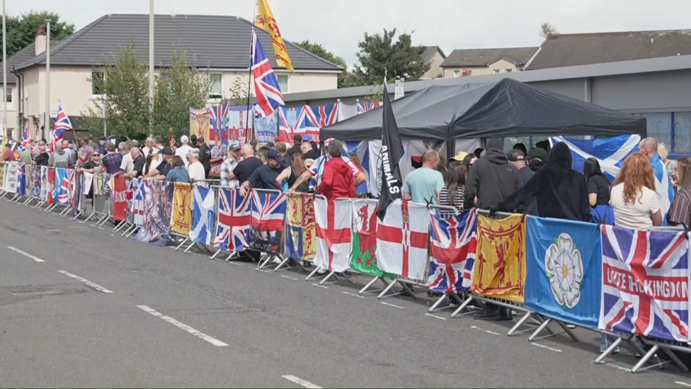 Anti-immigration protesters outside the Cladhan Hotel. They have numerous Union Jack, St George's and Lion Rampant flags draped over the railings they are standing behind and attched to a wall behind them. Others are flying above their heads.