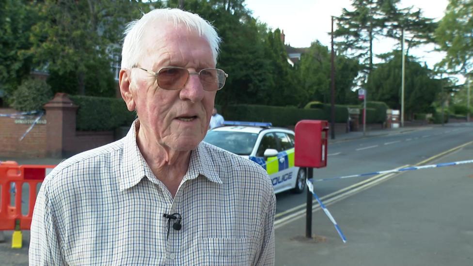 A man stands in the street with cordon and police tape behind him. He has white hair and wears glasses.