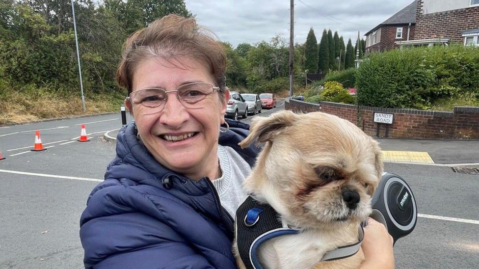 A woman in a navy puffer jacket and grey jumper smiles as she holds up a small dog in the street, with traffic cones across the road entrance.