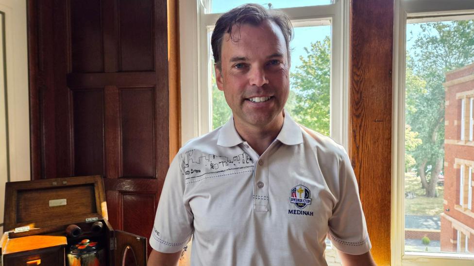 Ken Skates standing in front of a large window inside a wood-panelled room. The person is wearing a white polo shirt featuring the Ryder Cup 2012
