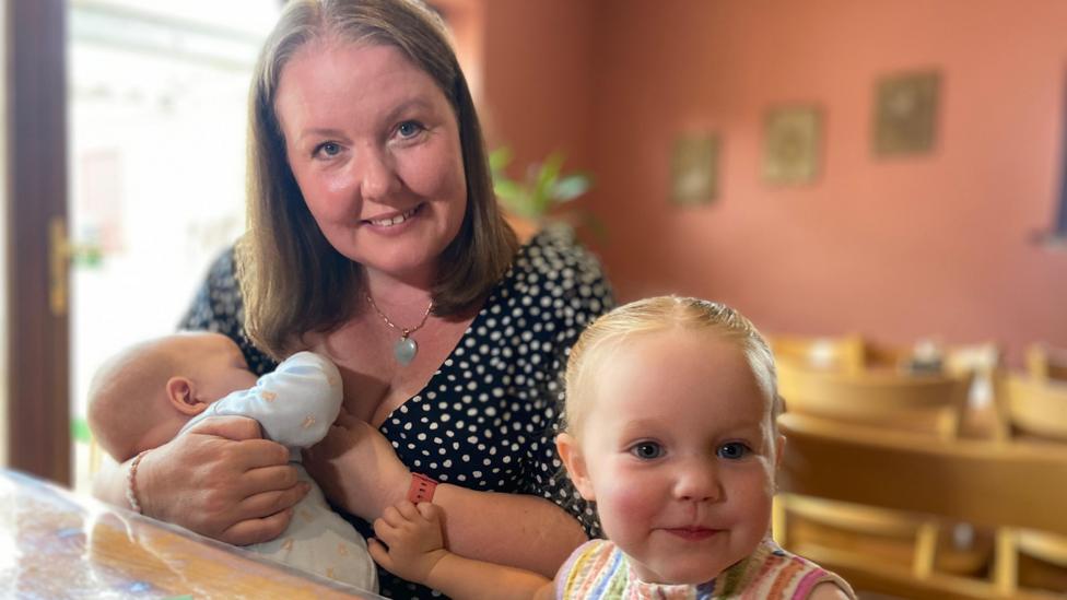 Katy, wearing a polka dot top and a chain with a light blue pendant, holds a small child chose to her chest, while another, slightly older child sits in front of them, wearing a colourful stripey top. 